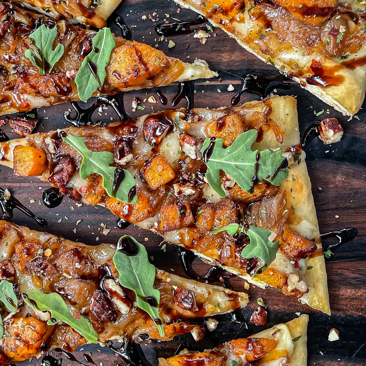 Enjoy the Butternut Squash & Caramelized Onion Flatbread (Kit)—flatbread slices topped with roasted vegetables, melted cheese, arugula, nuts, and a dark balsamic glaze drizzle, beautifully arranged on a wooden surface.